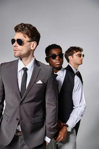 Modèles masculins multiraciaux attrayants dans des costumes élégants intelligents avec des lunettes de soleil posant sur fond gris — Photo de stock