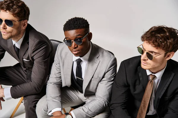 Attractive multicultural men with sunglasses in business attires sitting on chairs on gray backdrop — Stock Photo