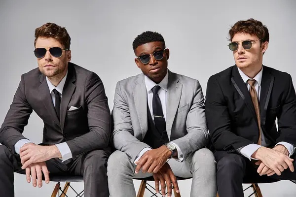 Good looking diverse men with sunglasses in business attires sitting on chairs on gray backdrop — Stock Photo