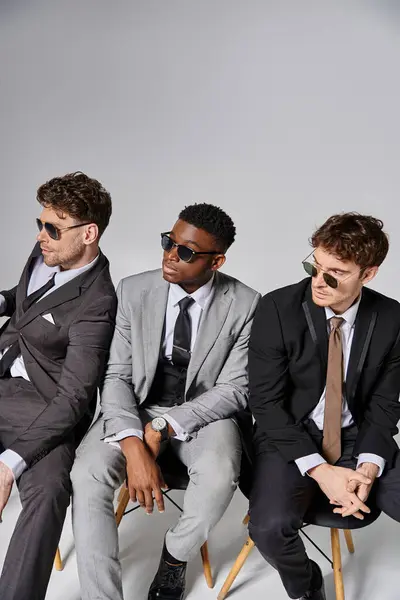 Handsome multiracial men with sunglasses in business attires sitting on chairs on gray backdrop — Stock Photo