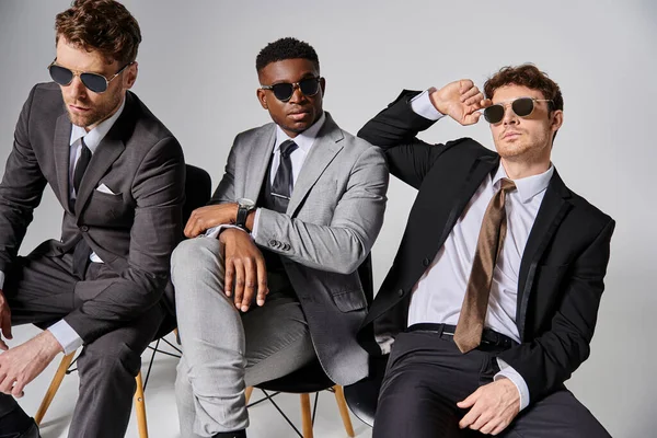 Homens inter-raciais bonitos com óculos de sol em trajes inteligentes sentados em cadeiras no fundo cinza — Fotografia de Stock