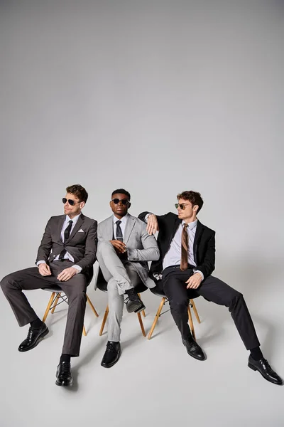 Homens inter-raciais atraentes com óculos de sol em trajes inteligentes sentados em cadeiras em pano de fundo cinza — Fotografia de Stock