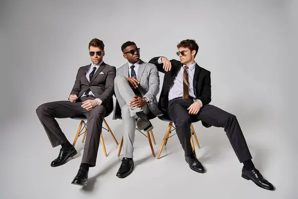 Atraentes homens inter-raciais com óculos de sol em trajes inteligentes sentados em cadeiras no fundo cinza — Fotografia de Stock