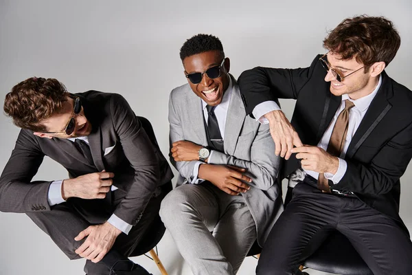 Happy young multicultural men in business smart suits sitting on chairs and smiling joyfully — Stock Photo