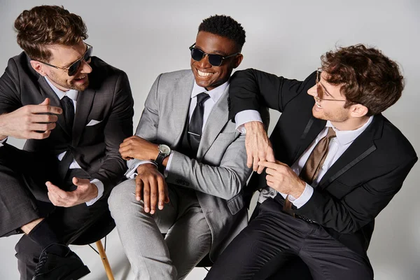 Homens multiculturais atraentes felizes em ternos inteligentes de negócios sentados em cadeiras e sorrindo alegremente — Fotografia de Stock
