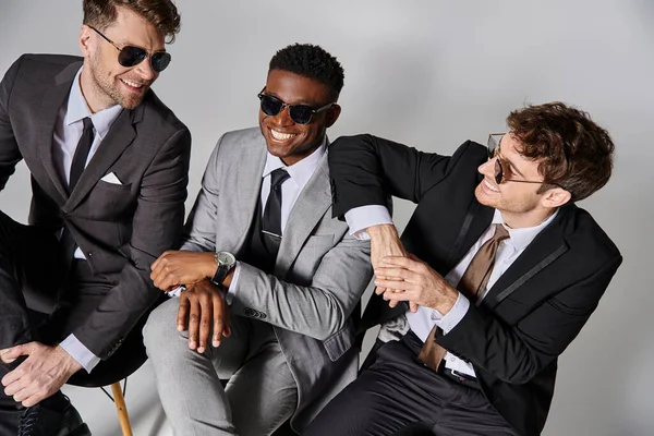 Happy good looking multicultural men in business smart suits sitting on chairs and smiling joyfully — Stock Photo