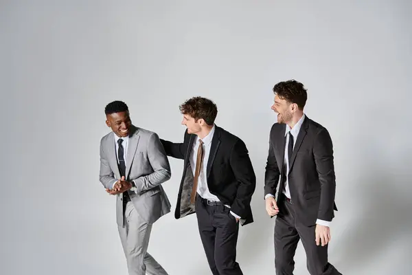 Amigos multiculturales alegres de moda en trajes elegantes de negocios sonriendo alegremente sobre fondo gris - foto de stock
