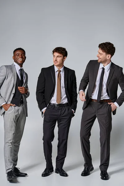 Добре виглядають веселі багаторасові чоловічі моделі в розумних костюмах, які щиро посміхаються на сірому тлі — стокове фото