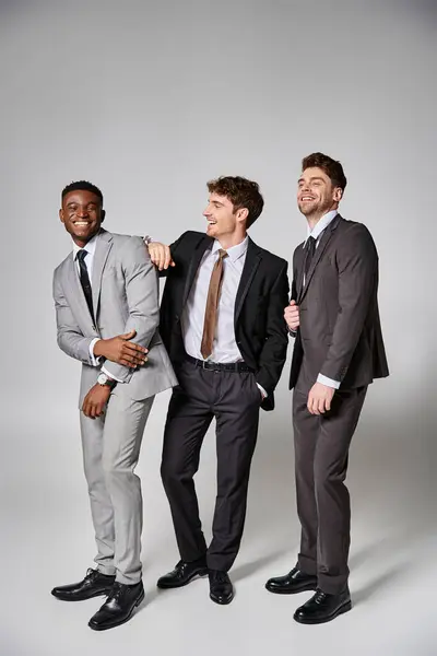 Good looking happy interracial male models in smart suits smiling sincerely on gray backdrop — Stock Photo