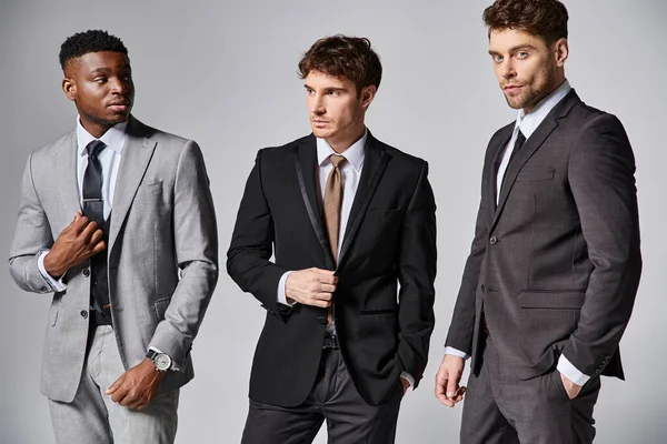Guapos modelos masculinos multiculturales con estilo en trajes elegantes de negocios posando sobre fondo gris - foto de stock