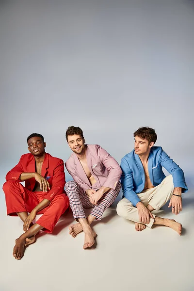 Joyous multiracial friends in vibrant colorful suits sitting on floor with crossed legs, fashion — Stock Photo