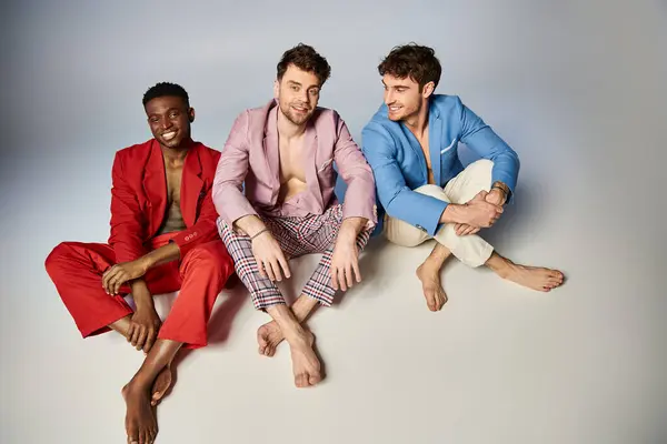 Amis interracial joyeux en costumes colorés vibrants assis sur le sol avec les jambes croisées, de la mode — Photo de stock