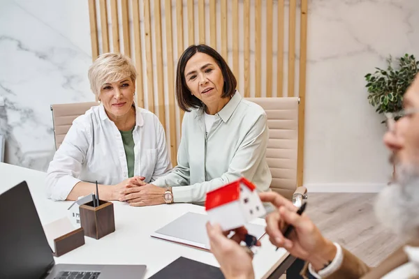 Middle aged interracial lgbt couple looking at realtor holding house model in real estate office — Stock Photo