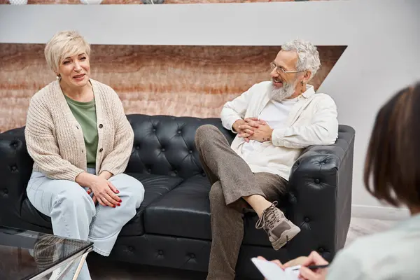 Happy middle aged couple sitting on leather and smiling with psychologist during consultation — Stock Photo