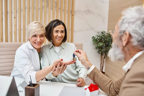 Middle aged realtor giving house key to happy interracial lgbt couple in real estate office — Stock Photo