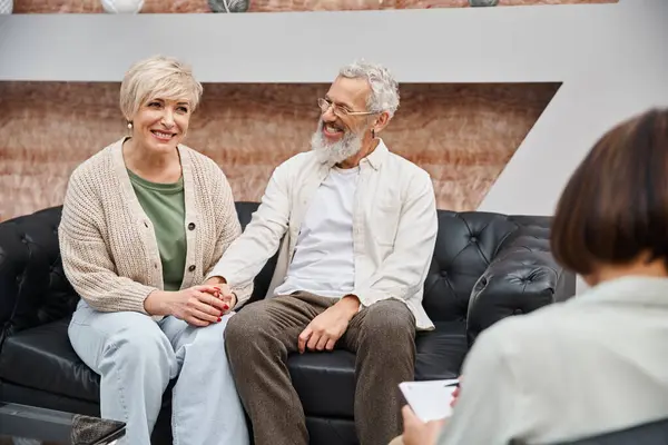 Heureux couple d'âge moyen assis sur le canapé en cuir et tenant la main près du conseiller familial — Photo de stock