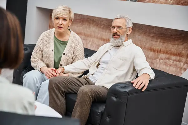 Middle aged couple sitting on leather couch and looking at psychologist during family consult — Stock Photo