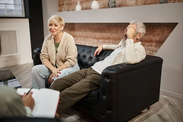 Alegre mujer de mediana edad sentada en el sofá de cuero cerca del marido durante la sesión de terapia familiar - foto de stock