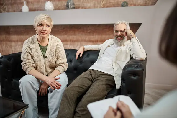 Mulher de meia idade sentada no sofá de couro perto do marido sorridente durante a sessão de terapia familiar — Fotografia de Stock