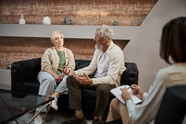 Бородатый мужчина разговаривает с женой средних лет, сидя рядом с психологом во время терапии — стоковое фото