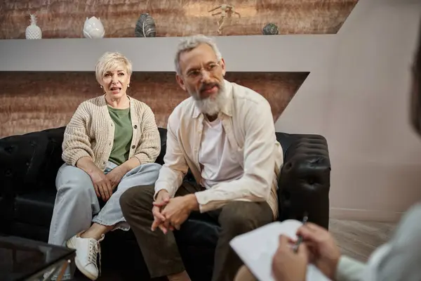 Casal de meia-idade emocional sentado no sofá e conversando com o psicólogo durante a sessão de terapia — Fotografia de Stock