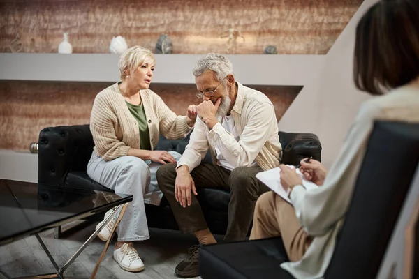 Cuidado esposa de mediana edad calmar marido barbudo durante la sesión de terapia familiar con el psicólogo - foto de stock