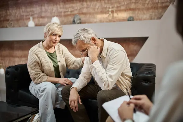 Турботлива дружина середнього віку заспокоює засмученого чоловіка під час сеансу сімейної терапії з психологом — стокове фото