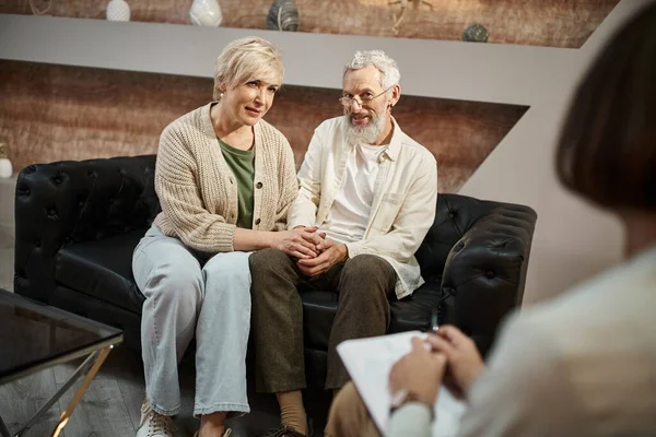 Подружня пара середнього віку тримає руки і сидить на шкіряному дивані під час сеансу сімейної терапії — стокове фото