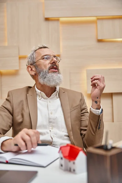 Tattooed middle aged realtor in glasses talking during consultation in real estate office — Stock Photo