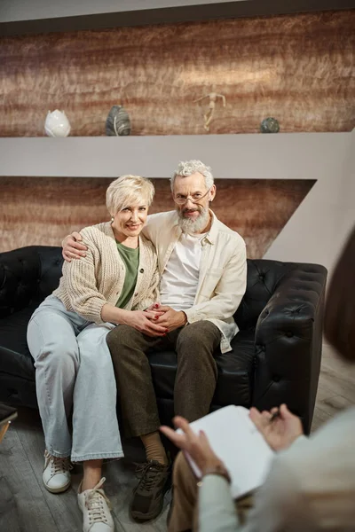 Feliz matrimonio de mediana edad pareja abrazando y sentado en el sofá de cuero durante la sesión de terapia familiar - foto de stock
