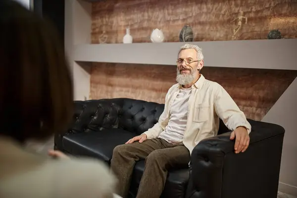 Hombre de mediana edad barbudo feliz con tatuaje sentado en el sofá de cuero y mirando al psicólogo - foto de stock