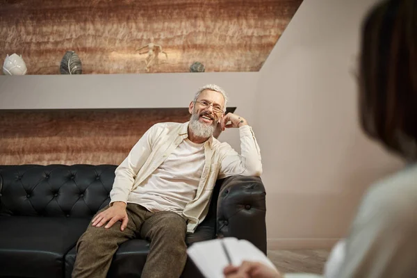 Cheerful bearded middled aged man with tattoo sitting on leather couch and looking at psychologist — Stock Photo