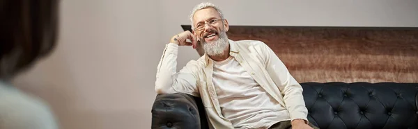 Alegre barbudo hombre de mediana edad sentado en el sofá de cuero y mirando psicólogo, pancarta - foto de stock