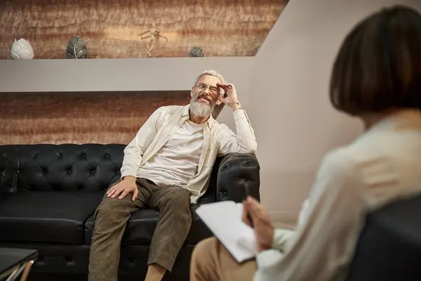 Cheerful bearded middled aged man with tattoo sitting on leather couch and looking at psychologist — Stock Photo