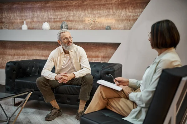 Homem de meia idade barbudo com tatuagem sentado no sofá de couro e falando com o psicólogo — Fotografia de Stock