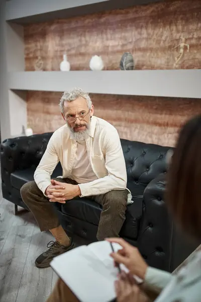 Bearded middled aged man with tattoo sitting on leather couch and listening to female psychologist — Stock Photo
