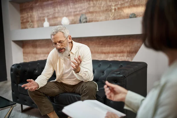 Worried bearded middled aged man with tattoo sitting on leather couch and talking to psychologist — Stock Photo