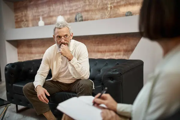 Worried bearded middled aged man with tattoo sitting on leather couch and looking at psychologist — Stock Photo
