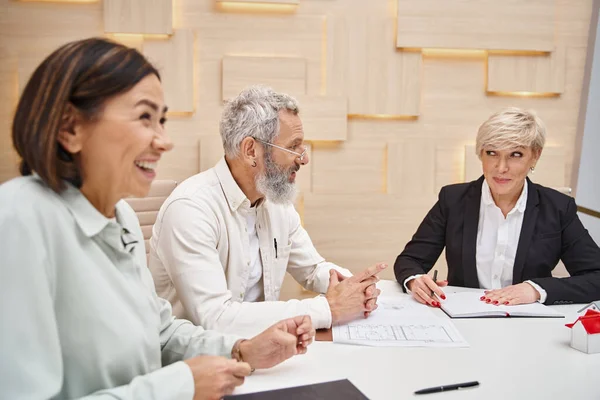 Happy middle aged man smiling near wife and female realtor while sitting near blueprint of new home — Stock Photo
