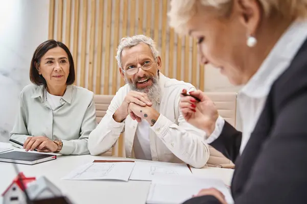 Happy middle aged married couple smiling near female realtor and sitting near blueprint of new home — Stock Photo