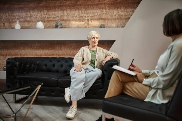 Focus on middled aged blonde woman sitting on couch and talking to psychologist during session — Stock Photo