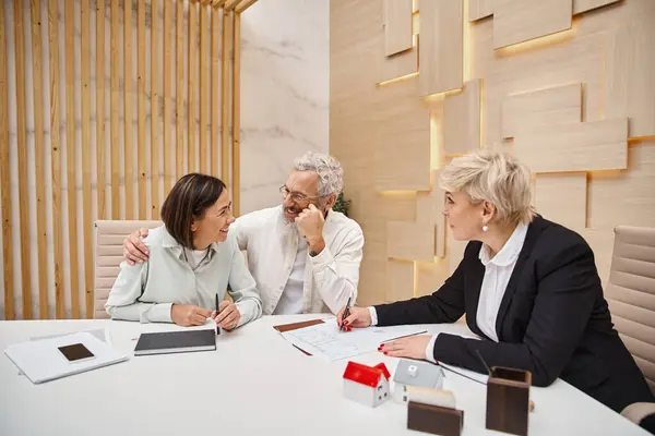 Cheerful middle aged married couple smiling near female realtor and discussing project of new home — Stock Photo