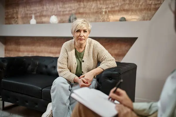 Focus on blonde middled aged woman sitting on couch near psychologist taking notes during session — Stock Photo