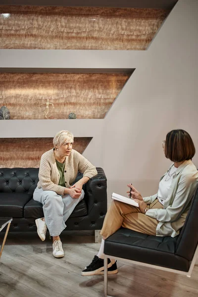 Блондинка среднего возраста, сидящая на диване и разговаривающая с психологом во время сеанса терапии — стоковое фото