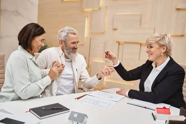 Agente inmobiliario de mediana edad dando la llave de la casa nueva a la pareja casada feliz en la oficina de bienes raíces - foto de stock