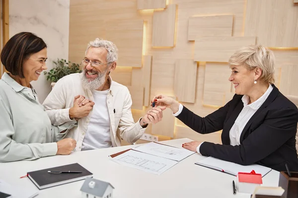 Makler mittleren Alters übergibt Schlüssel für neues Haus an glückliches Ehepaar im Immobilienbüro — Stockfoto