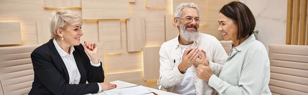 Middle aged realtor looking at happy married couple holding key from new house in office, banner — Stock Photo