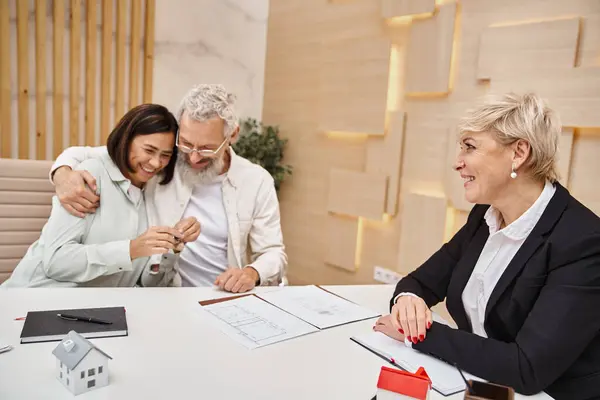 Cheerful married couple holding key from new house near smiling realtor in real estate office — Stock Photo