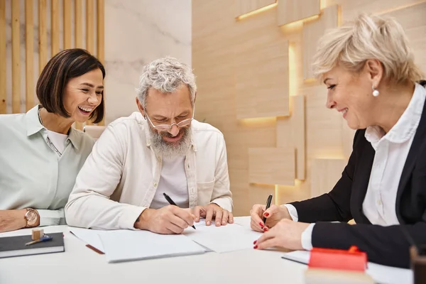 Счастливый мужчина средних лет, подписывающий контракт рядом с женой и счастливым агентом по недвижимости в офисе, новый дом — стоковое фото