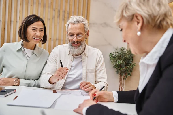 Happy middle aged realtor signing contact near married couple in real estate office, selling house — Stock Photo